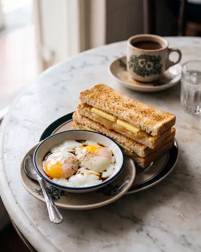 Kaya toast: crisp thin toast sandwiching a thick layer of pale-golden coconut-egg kaya and a cold slab of butter, cut into fingers, beside two soft-boiled eggs with runny yolks in a bowl with dark soy and white pepper.